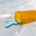 An overturned orange prescription bottle with blue tablets spilled out on a light blue background.