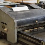 An old printing press with a blank sheet of paper on it in a workshop setting.
